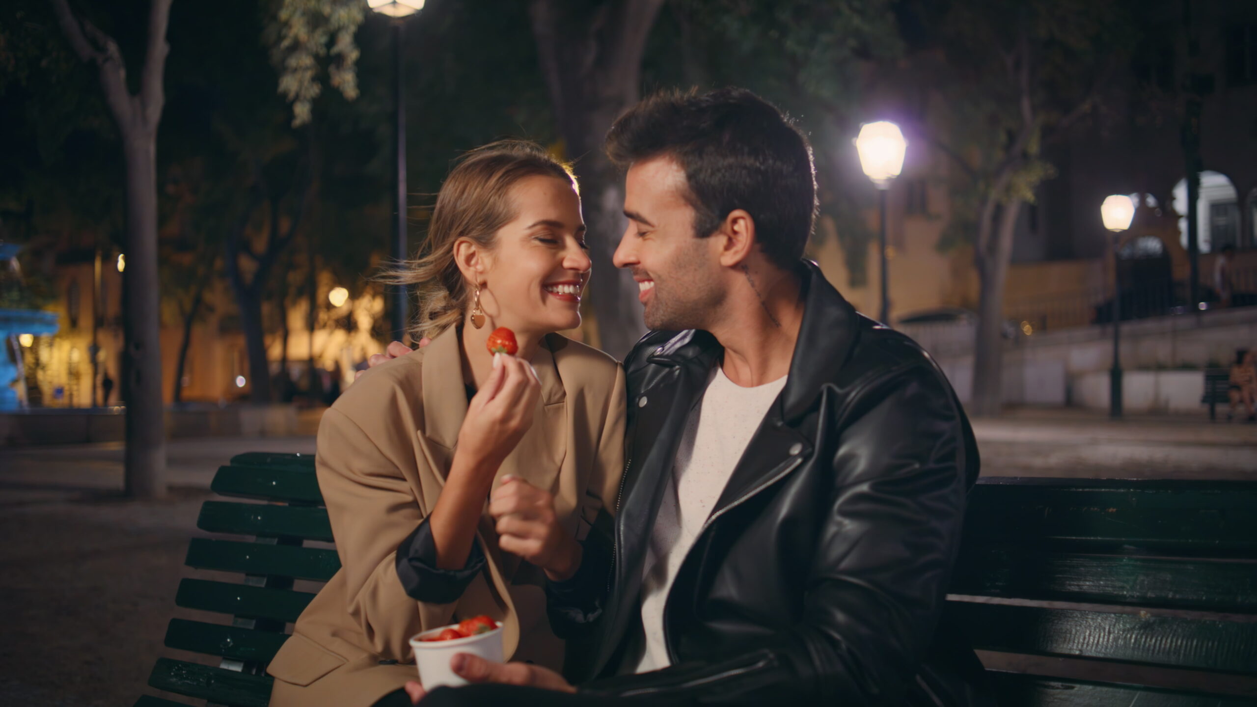 Relaxed partners eating strawberry in city park. Beautiful girl enjoying romantic date tasting sweet berry in evening. Handsome hispanic guy embracing girlfriend. Affectionate married people have fun